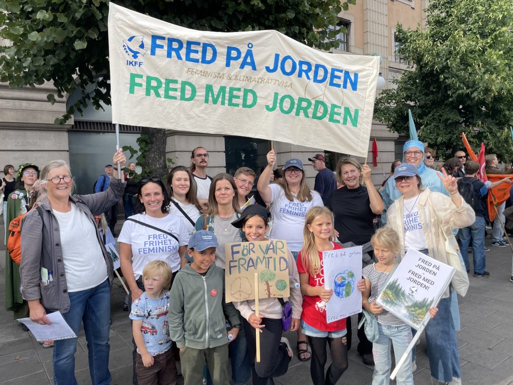 Ett gäng IKFF-are, vuxna och barn, med en banderoll där det står Fred på jorden, fred med jorden - feminism och klimaträttvisa.